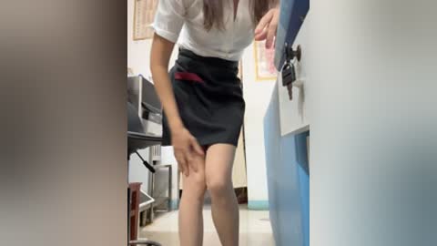 A video captures a woman in a white blouse and black skirt, bending slightly, in a hallway with light blue walls and office equipment.