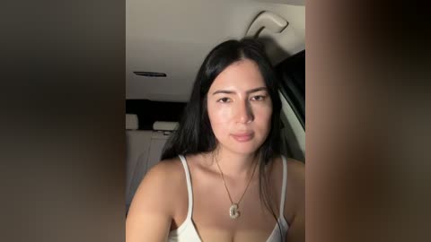 Video of a young woman with long black hair, wearing a white spaghetti-strap top, standing indoors near a car, with a blurred background.