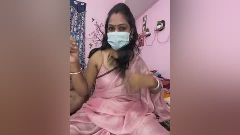 A video of an Indian woman with medium skin tone, wearing a pink saree, face mask, and bracelets, taking a selfie in a pink room with a floral-patterned wall hanging and framed photos.