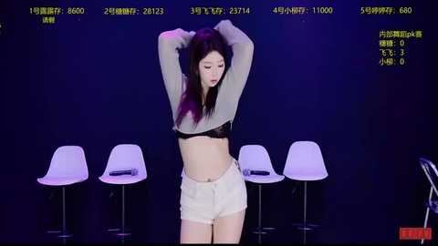 A video of a young Asian woman with long dark hair, wearing a gray crop top and white shorts, posing in front of three white stools against a dark background.