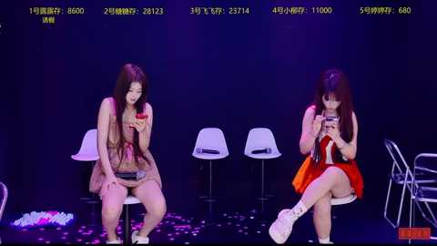 Video of two Asian women with long hair, seated on white chairs, texting, in a dimly lit room with abstract purple and blue lighting.