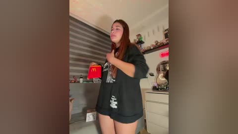 Video of a young woman with long, reddish-brown hair, wearing a black oversized shirt, standing in a brightly lit, cluttered room with a McDonald's bag and toys visible in the background.