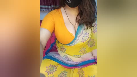 Video of a woman with light brown skin, wearing a yellow sari with blue and pink floral patterns, and a black face mask, seated on a bed with colorful bedding.