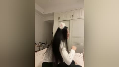 Video of a young Asian woman with long black hair, wearing a white maid outfit, seated on a bed in a dimly lit bedroom with white cabinets and a cluttered nightstand.