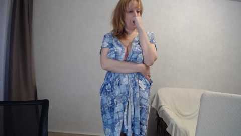A video shows a middle-aged woman with light skin and shoulder-length blonde hair, wearing a blue floral patterned dress, covering her mouth in a beige room with a white couch.