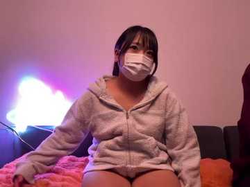 Video of an Asian woman with straight black hair, wearing a white surgical mask, gray hoodie, and pink panties, sitting on a bed with a pink blanket. Background includes a purple light and a maroon curtain.