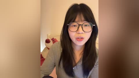 Video of an Asian woman with straight black hair and glasses, wearing a gray hoodie, standing in a bedroom with beige walls and a teddy bear on a bed.