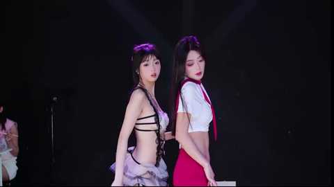 Video of two East Asian women, one in a black strappy top, the other in a white crop top and red suspenders, against a dark stage backdrop.