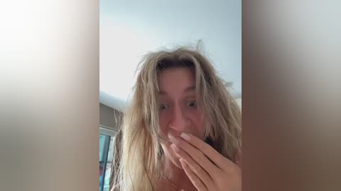 Video of a blonde woman with messy hair, covering her mouth in a playful manner, standing indoors near a window with soft lighting.