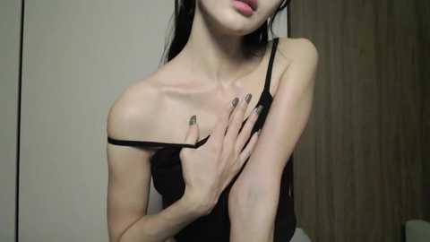 Video of an Asian woman with fair skin, long black hair, and full lips. She's wearing a black spaghetti strap top, her left hand covering her chest, revealing a slender, petite physique. The background is a wooden door and a beige wall.