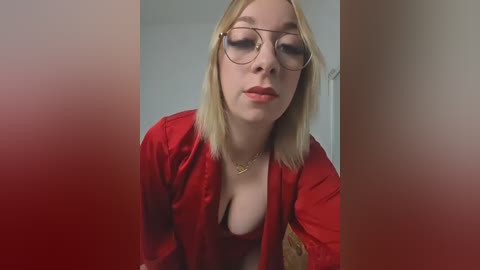 A video of a young Caucasian woman with light skin, straight blonde hair, and glasses, wearing a red satin robe with a black lace bra, leaning forward.