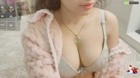 A video of an Asian woman with long black hair, wearing a light pink fluffy robe, revealing a white bra that highlights her ample cleavage. She's indoors, likely in a bedroom, with a white bed and a partially visible wall.