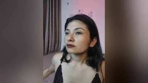 A video of an Asian woman with long black hair, wearing a black lace bra, in a dimly lit room with a pinkish hue and a curtain in the background.