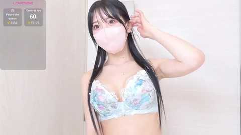 Video of an East Asian woman with long black hair and light skin, wearing a pastel floral bra and a pink face mask. She's indoors, holding her hair up with one hand.
