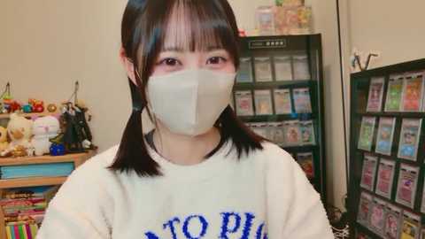 Video of a young Asian woman with black pigtails wearing a white T-shirt, a face mask, and a surgical mask, in a toy store with shelves of toys, stuffed animals, and games in the background.