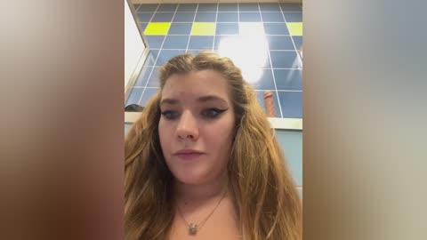 Video of a young woman with long, wavy blonde hair, light skin, and full lips, wearing black eyeliner and a gold necklace. She stands in a tiled bathroom with yellow and blue checkered walls.
