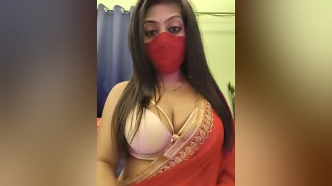 Media: Video of a South Asian woman with medium skin tone, wearing a red sari, beige bra, and red face mask, standing in a bathroom with blue curtains and white tiles.
