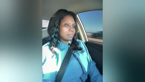 Video of a woman with medium skin tone and shoulder-length, wavy black hair, wearing a light blue shirt and a black seatbelt, sitting in a car with a blue sky visible through the window.