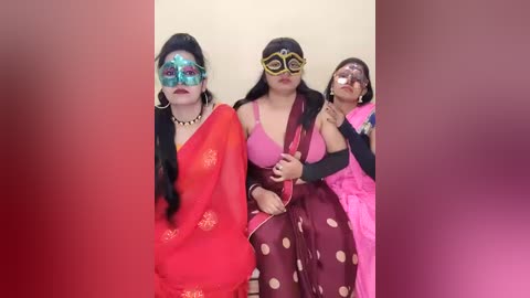 Video of three women in colorful, traditional Indian attire, adorned with elaborate, vibrant masks and accessories, set against a blurred background of red and pink hues.