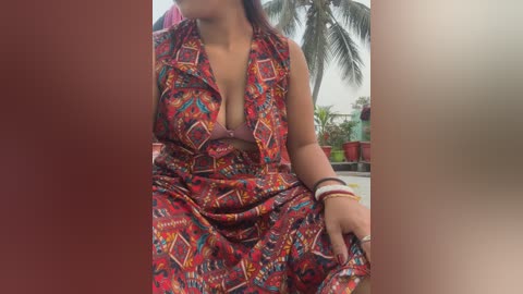 Video of a woman with medium brown skin, wearing a vibrant red and orange patterned sari, seated outdoors near a palm tree and potted plants.