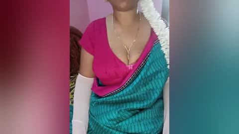 A video of a woman wearing a vibrant pink top, a teal and white striped sari, and white elbow-length gloves. Her neck is adorned with a gold necklace. The background features a pink wall and a floral-patterned cushion.