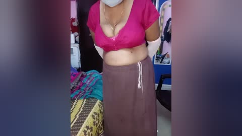 A video shows a woman wearing a pink crop top, exposing her midriff, and a brown skirt, standing in a cluttered bedroom.