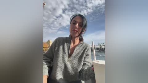 Video of a woman with medium-length dark hair, wearing a gray, textured cardigan, standing on a boat deck with a cloudy sky and a yellow building in the background.