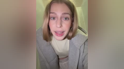 Video of a young, fair-skinned girl with straight, shoulder-length blonde hair, wearing a gray coat over a beige turtleneck. She has braces and is smiling, with a soft, blurred background.