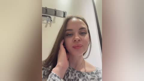 A video of a young Caucasian woman with fair skin and long brown hair, wearing a leopard-print top, leaning against a white wall with a metallic hook and a blurred mirror reflection.