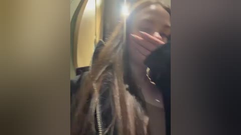 Video of a young woman with long, blonde hair, wearing a dark jacket, partially hiding her face with her hand, in a dimly lit room with beige walls.