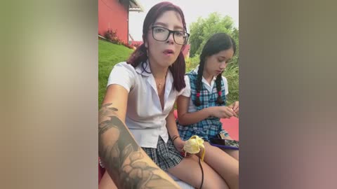 Video of two young girls, one with glasses and a tattooed arm, in school uniforms, sitting on a grassy lawn beside a red building.