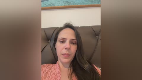 A video shows a middle-aged woman with long dark hair, light skin, and a serious expression, wearing a pink lace blouse. She is seated on a brown tufted leather couch in a room with a turquoise rug and framed artwork on the wall.