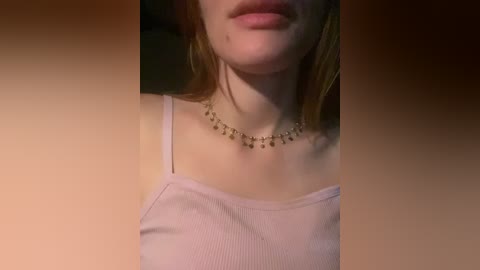 A video of a young woman with fair skin and shoulder-length auburn hair, wearing a light pink spaghetti-strap top and a delicate, beaded necklace. The background is blurred, with warm lighting highlighting her face and upper body.