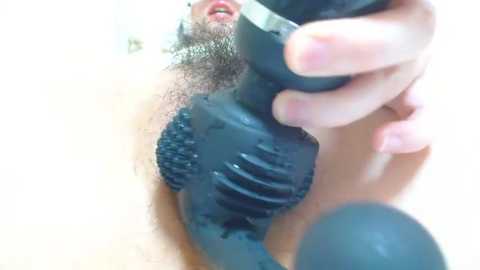 A close-up video shows a person's hand using a black vibrating wand on their pubic area, revealing a trimmed pubic hair. The background is blurred and white.