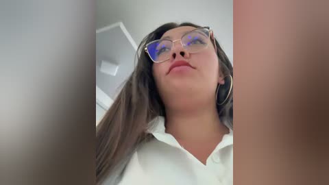 A close-up video of a young Asian woman with long dark hair, wearing glasses and a white collared shirt. The background is blurry with white walls and a ceiling light.