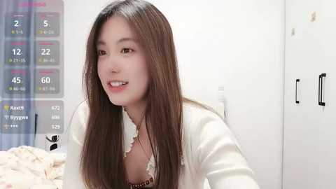 Video of a young East Asian woman with long, straight brown hair, wearing a white blouse, smiling. Background features a white wall, a chart with weather data, and a window with a blue curtain.
