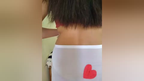 Video of a woman's back in a red bra, wearing a white skirt with a large, embroidered red heart on the left side. The background is blurred, showing a white wall.