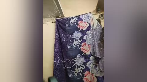 A video of a shower curtain with a dark purple background adorned with swirling white patterns and large, detailed pink and white roses. The curtain hangs in a plain, beige-tiled bathroom.