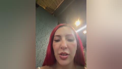 A video of a young woman with fair skin, red hijab, and a slightly open mouth, taken indoors with a textured teal wall and wooden ceiling beams in the background.