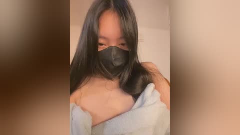 Video of an Asian woman with long black hair, wearing a black face mask, partially covering her chest with a light gray towel.