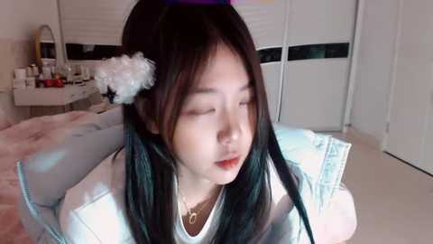 Video of an East Asian woman with long black hair, wearing a white shirt, and a white flower headband, sitting on a bed in a brightly lit bedroom with white walls and a vanity table.