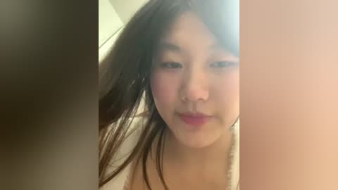 Video of an East Asian woman with long black hair, fair skin, and pink lips, wearing a white top, standing in front of a blurred beige wall.