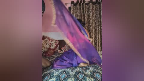 A video of a woman with brown skin and long, dark hair, wearing a flowing purple and pink dress, seated on a patterned bedspread. She is partially obscured by the fabric. The background features floral curtains and a wall with red and white tiles.