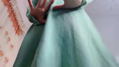 A video captures a person wearing a light green hospital gown, with their left hand pressing against their abdomen. The background features a partially visible floral-patterned curtain on the left. The image is taken from a low angle, emphasizing the gown's texture and the person's hand.