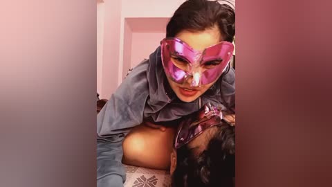 Video of a woman in a reflective pink mask and blue robe, with a visible exposed breast, leaning over a man with dark hair, in a dimly lit room.