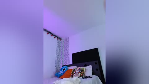 Video of a minimalist bedroom with a black headboard, white sheets, and a blue plush toy. The room is softly lit with purple and white lighting.