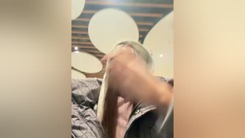 A blurred video of a blonde woman with shoulder-length hair, wearing a gray jacket, standing against a background with round, white, decorative lights.