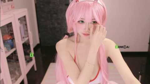 Video of a slender, fair-skinned woman with pink hair and green eyes, wearing a revealing, strapless, pink dress, sitting on a white floor. Background includes white shelves and a black door.