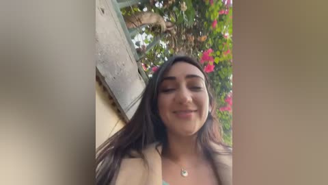 Video of a young woman with long dark hair, wearing a beige top, smiling, against a backdrop of lush greenery and vibrant pink flowers.