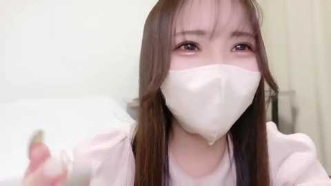 A video of an Asian woman with long, straight brown hair, wearing a white surgical mask and light-colored scrubs, holding a syringe in her right hand. She has a serious expression. The background is a blurred medical setting.